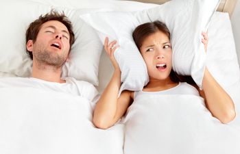 A snoring man sleeping next to an annoyed woman covering her ears with a pillow.